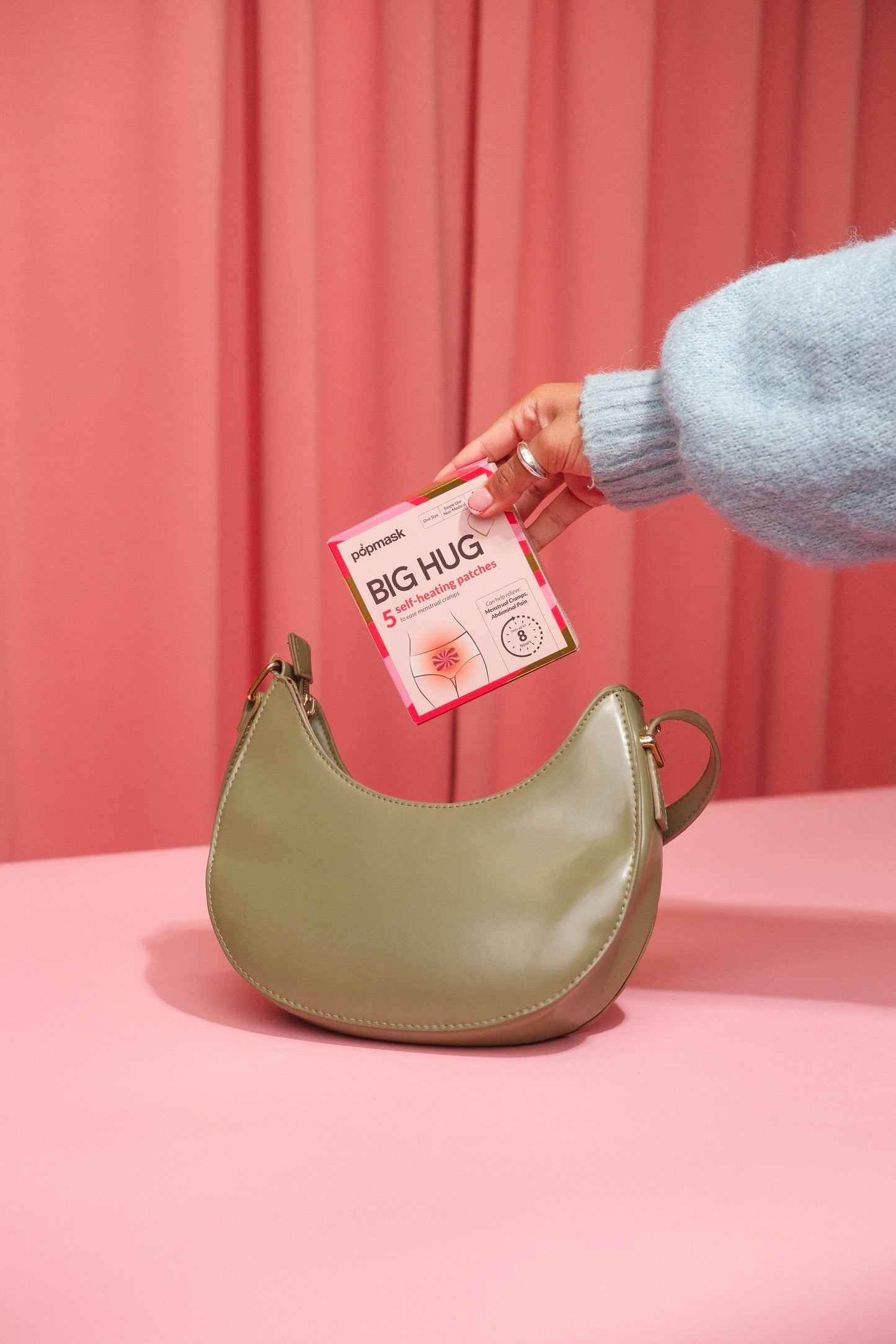 Person holding a green handbag with a pink product labeled 'Big Hug' against a pink background